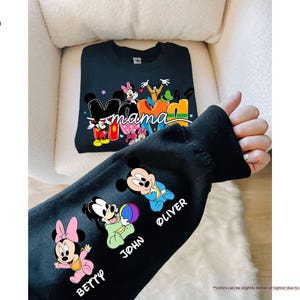 K&ouml;nnte beinhalten: Schwarzes Sweatshirt mit einem farbenfrohen "Mama"-Design mit Mickey- und Minnie-Mouse-Figuren. Der &Auml;rmel zeigt Cartoon-Bilder von Minnie, Mickey und zwei weiteren Figuren mit den Namen "Betty", "John" und "Oliver".