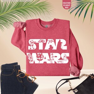 May include: Heather scarlet red sweatshirt with the text "STAR WARS" in white block letters. The sweatshirt features white illustrations of characters and vehicles from the Star Wars franchise. The long-sleeved shirt is paired with blue jeans and a black purse.