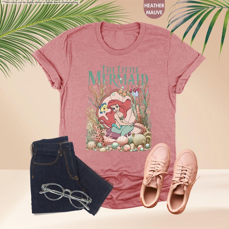 May include: Heather mauve t-shirt featuring "The Little Mermaid" graphic. The design includes Ariel, Flounder, and other sea creatures. Paired with dark blue jeans, glasses, and pink sneakers. The shirt is made of a soft material.