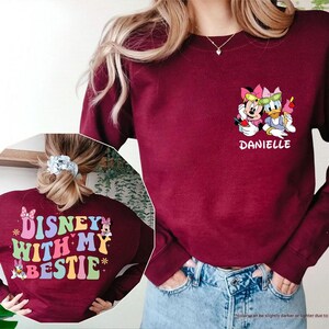 May include: A maroon long-sleeved sweatshirt with a colorful graphic on the back that says "DISNEY WITH MY BESTIE" and a graphic of Minnie Mouse and Daisy Duck on the front with the name "DANIELLE" underneath.