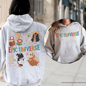 May include: A light gray hoodie with the text "EPIC UNIVERSE" in colorful letters. The back of the hoodie features various cartoon characters and the same text. The hoodie is made of a soft material and has a hood and a front pocket.