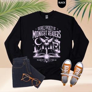 May include: Black sweatshirt with the text "SECRET SOCIETY OF MIDNIGHT READERS" and an owl graphic. The design includes a crescent moon, candles, and an hourglass. The sweatshirt is paired with blue jeans and orange sneakers.