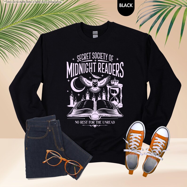 May include: Black sweatshirt with the text "SECRET SOCIETY OF MIDNIGHT READERS" and an owl graphic. The design includes a crescent moon, candles, and an hourglass. The sweatshirt is paired with blue jeans and orange sneakers.