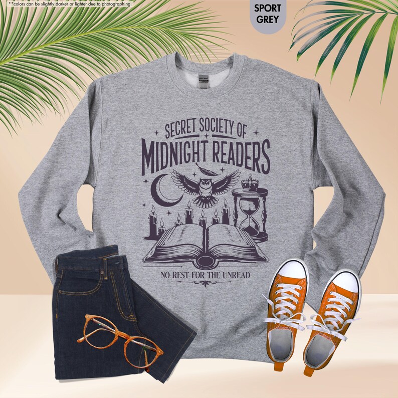 May include: A gray sweatshirt with the text "SECRET SOCIETY OF MIDNIGHT READERS" and an owl graphic, paired with dark blue jeans, orange-framed glasses, and orange sneakers. The sweatshirt design includes a crescent moon, candles, and an hourglass.
