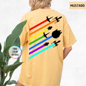 K&ouml;nnte beinhalten: Ein senfgelbes T-Shirt mit einem Regenbogenmuster und Silhouetten von Star Wars Raumschiffen. Der Text "MUSTARD" befindet sich in der oberen rechten Ecke des Bildes.