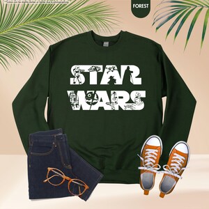 May include: A forest green sweatshirt with the words "STAR WARS" in white, featuring character silhouettes. The image includes a pair of dark wash jeans, orange sneakers, and eyeglasses.