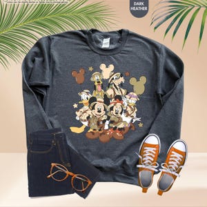 May include: Dark heather gray sweatshirt featuring a graphic of Disney characters in safari outfits. The image includes Mickey and Minnie Mouse, Donald Duck, Goofy, and others. The sweatshirt is paired with dark wash jeans, orange sneakers, and tortoiseshell glasses.
