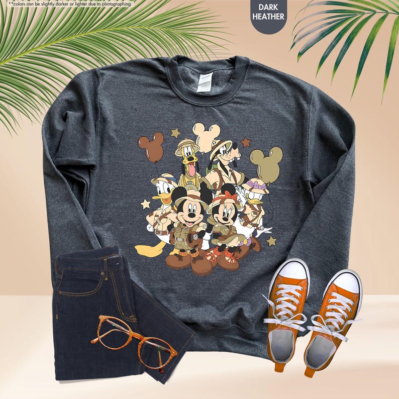May include: Dark heather gray sweatshirt featuring a graphic of Disney characters in safari outfits. The image includes Mickey and Minnie Mouse, Donald Duck, Goofy, and others. The sweatshirt is paired with dark wash jeans, orange sneakers, and tortoiseshell glasses.