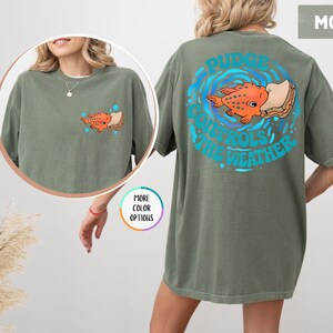 Puede incluir: Camiseta verde musgo con un dise&ntilde;o de pez de dibujos animados. La parte delantera de la camiseta presenta una peque&ntilde;a ilustraci&oacute;n de pez, mientras que la parte trasera muestra un dise&ntilde;o m&aacute;s grande con el texto "Pudge Controls The Weather" en azul. La camiseta es de corte holgado.