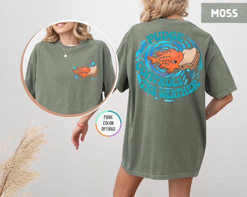 Puede incluir: Camiseta verde musgo con un dise&ntilde;o de pez de dibujos animados. La parte delantera de la camiseta presenta una peque&ntilde;a ilustraci&oacute;n de pez, mientras que la parte trasera muestra un dise&ntilde;o m&aacute;s grande con el texto "Pudge Controls The Weather" en azul. La camiseta es de corte holgado.