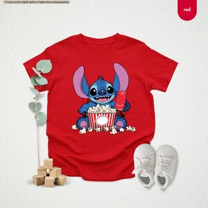 May include: A red t-shirt featuring a cartoon character holding popcorn and a drink. The character is blue with large ears and is sitting in front of a red and white striped popcorn container. White baby shoes are next to the shirt.