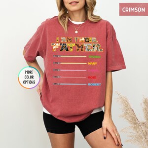 Puede incluir: Camiseta color carmes&iacute; con el texto "I AM THE FATHER" en un dise&ntilde;o de patchwork. Debajo, sables de luz coloridos con los nombres: Thomas, Mary, Daniel, Rose y Robert. La camiseta tiene un corte holgado.