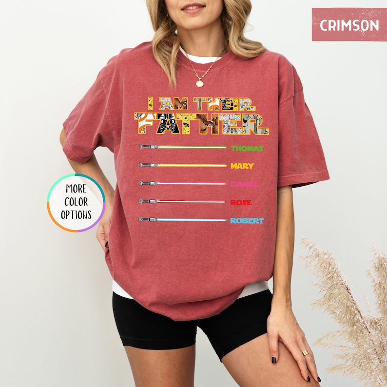 Puede incluir: Camiseta color carmes&iacute; con el texto "I AM THE FATHER" en un dise&ntilde;o de patchwork. Debajo, sables de luz coloridos con los nombres: Thomas, Mary, Daniel, Rose y Robert. La camiseta tiene un corte holgado.