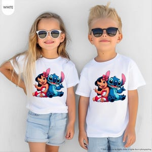 K&ouml;nnte beinhalten: Zwei Kinder tragen wei&szlig;e T-Shirts mit einer farbenfrohen Grafik von Lilo und Stitch. Die Zeichentrickfiguren sind in einer freundlichen Umarmung dargestellt. Die Shirts werden mit hellblauen Jeansshorts kombiniert.