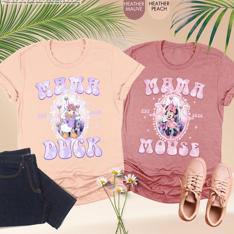 May include: Two heather peach and mauve t-shirts with the word "MAMA" in a bubble font. One shirt features Daisy Duck, the other Minnie Mouse, both in a decorative frame. The shirts are next to a pair of jeans and sneakers.