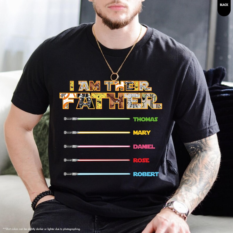 Puede incluir: Camiseta negra con el texto "I AM THEIR FATHER" en amarillo y blanco, con nombres y dise&ntilde;os de sables de luz debajo. Se enumeran los nombres Thomas, Mary, Daniel, Rose y Robert. La camiseta es de manga corta.