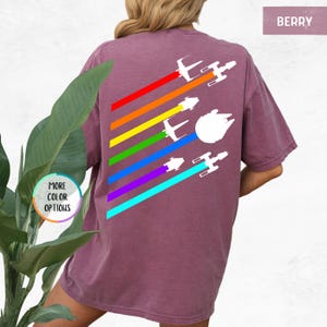 K&ouml;nnte beinhalten: Ein lila T-Shirt mit einem Regenbogenmuster und wei&szlig;en Silhouetten von Star Wars Raumschiffen. Der Text "BERRY" befindet sich in der oberen rechten Ecke des Bildes.