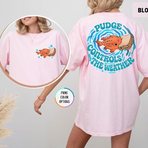 Puede incluir: Camiseta rosa claro con un dise&ntilde;o de pez naranja y s&aacute;ndwich, con el texto "Pudge Controls The Weather" en la espalda. La camiseta tiene un peque&ntilde;o gr&aacute;fico en la parte delantera. La palabra "Blossom" est&aacute; en la esquina superior derecha.