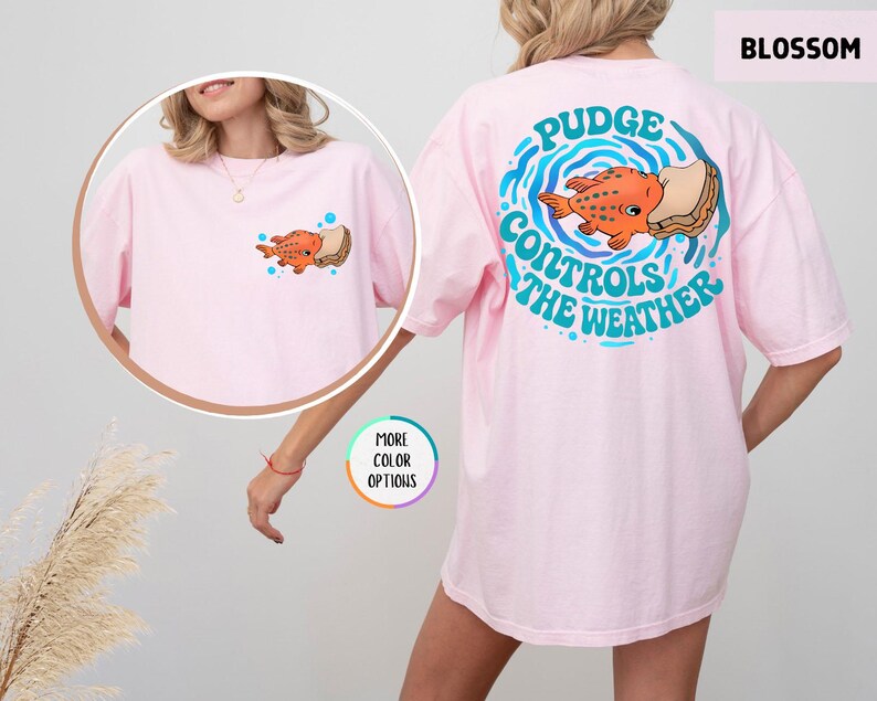 Puede incluir: Camiseta rosa claro con un dise&ntilde;o de pez naranja y s&aacute;ndwich, con el texto "Pudge Controls The Weather" en la espalda. La camiseta tiene un peque&ntilde;o gr&aacute;fico en la parte delantera. La palabra "Blossom" est&aacute; en la esquina superior derecha.