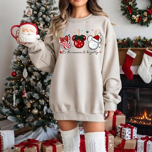 May include: A beige sweatshirt with a festive design featuring candy cane, red bow, and Mickey Mouse ornament graphics. The text "It's the season to be jolly" is printed below the ornaments. A Santa mug is held in the hand.