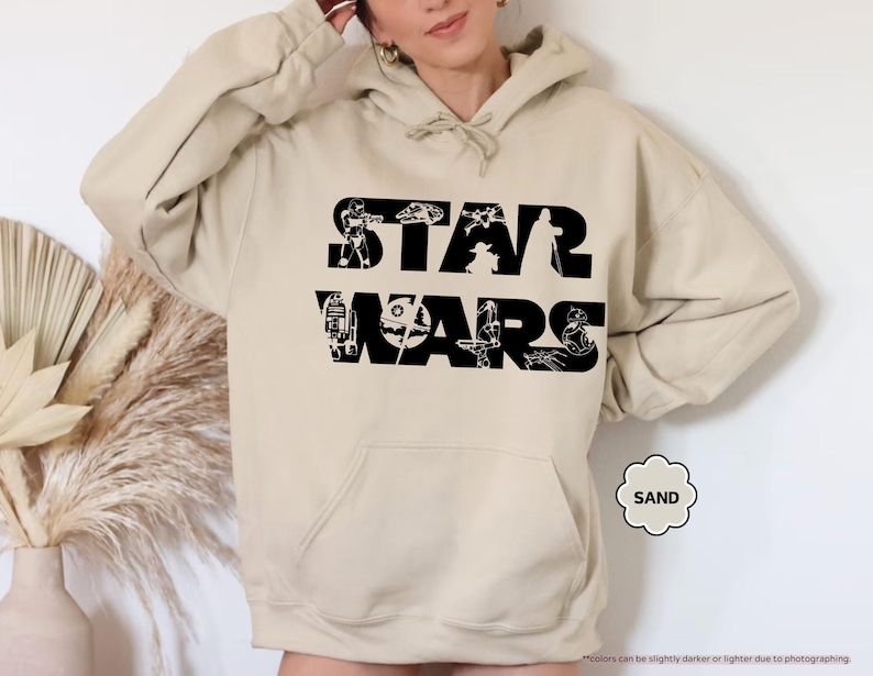 May include: Sand-colored hoodie featuring the Star Wars logo in black. The logo incorporates various character silhouettes within the letters. The hoodie has a front pocket and a drawstring hood. The word "SAND" is in a small, decorative circle.
