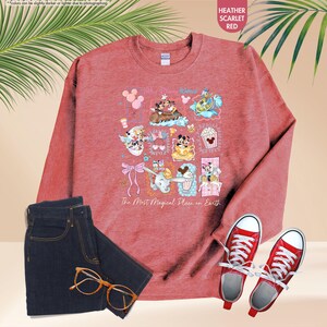 May include: Heather scarlet red sweatshirt with a colorful graphic of Disney characters and the text "The Most Magical Place on Earth." The image also includes dark blue jeans, red sneakers, and gold-rimmed glasses.