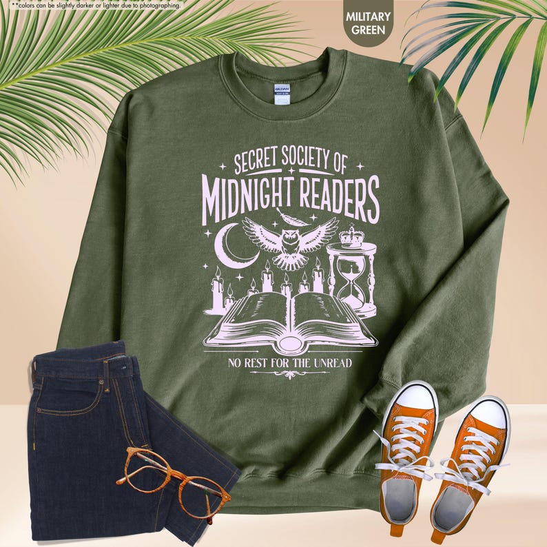 May include: Military green sweatshirt with the text "SECRET SOCIETY OF MIDNIGHT READERS" and a graphic of an owl, moon, candles, and an open book. The sweatshirt is paired with dark blue jeans, orange sneakers, and glasses.