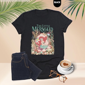 May include: Black t-shirt featuring a colorful graphic of Ariel from The Little Mermaid, with the text "The Little Mermaid". The design includes coral, shells, and other sea life. A pair of dark blue jeans, a cup of coffee, and glasses are also in the image.
