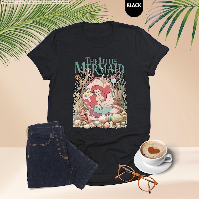 May include: Black t-shirt featuring a colorful graphic of Ariel from The Little Mermaid, with the text "The Little Mermaid". The design includes coral, shells, and other sea life. A pair of dark blue jeans, a cup of coffee, and glasses are also in the image.