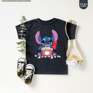 May include: Dark grey heather t-shirt featuring a cartoon character holding a red soda and a popcorn bucket. The character is blue with large ears and is sitting in a pile of popcorn. The shirt has a crew neck and short sleeves.