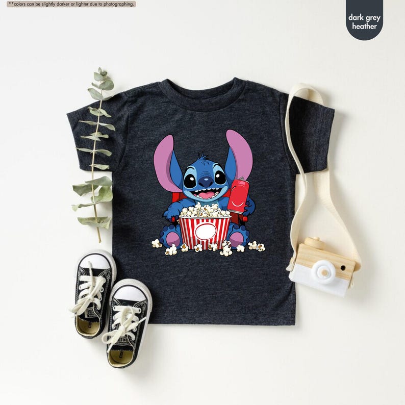May include: Dark grey heather t-shirt featuring a cartoon character holding a red soda and a popcorn bucket. The character is blue with large ears and is sitting in a pile of popcorn. The shirt has a crew neck and short sleeves.