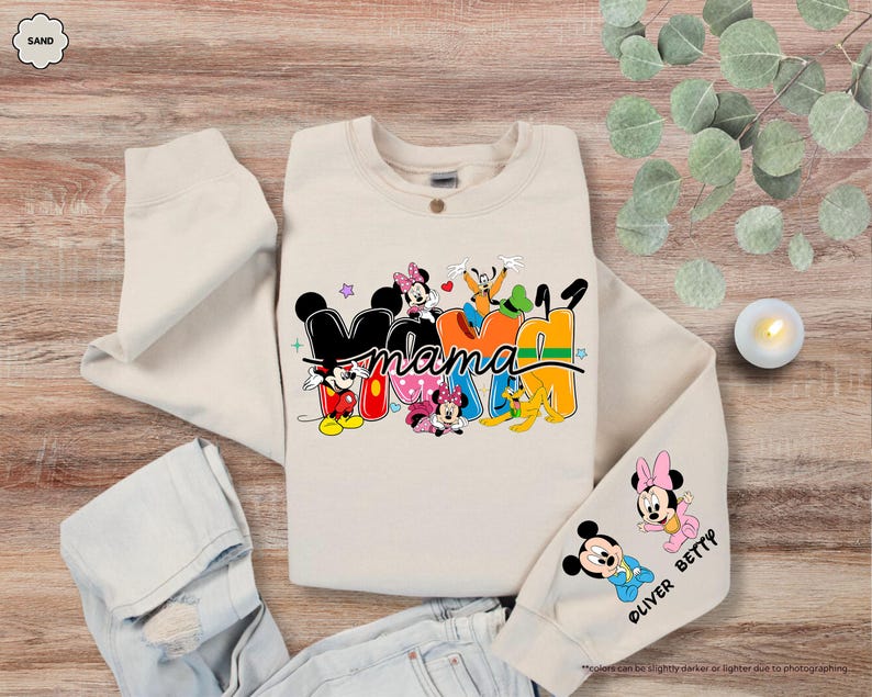 K&ouml;nnte beinhalten: Ein sandfarbenes Sweatshirt mit dem Wort "Mama" in einem farbenfrohen Disney-Design. Das Design umfasst Micky und Minnie Maus, Goofy und Pluto. Der &Auml;rmel hat zus&auml;tzliche Minnie- und Micky-Maus-Grafiken mit dem Text "OLIVER BETTY".