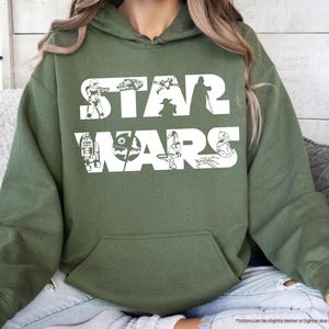 May include: Military green hoodie with a white "STAR WARS" graphic. The graphic features iconic characters and elements from the Star Wars franchise. The hoodie has a front pocket and a drawstring hood.