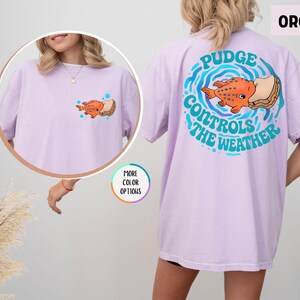 Puede incluir: Camiseta color orqu&iacute;dea con un dise&ntilde;o de pez de dibujos animados y s&aacute;ndwich. La parte trasera de la camiseta tiene un dise&ntilde;o circular con el texto "Pudge Controls The Weather". La parte delantera tiene una versi&oacute;n m&aacute;s peque&ntilde;a del gr&aacute;fico del pez.