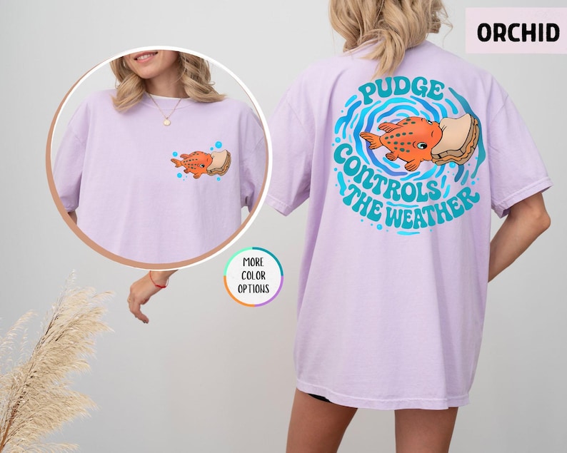 Puede incluir: Camiseta color orqu&iacute;dea con un dise&ntilde;o de pez de dibujos animados y s&aacute;ndwich. La parte trasera de la camiseta tiene un dise&ntilde;o circular con el texto "Pudge Controls The Weather". La parte delantera tiene una versi&oacute;n m&aacute;s peque&ntilde;a del gr&aacute;fico del pez.