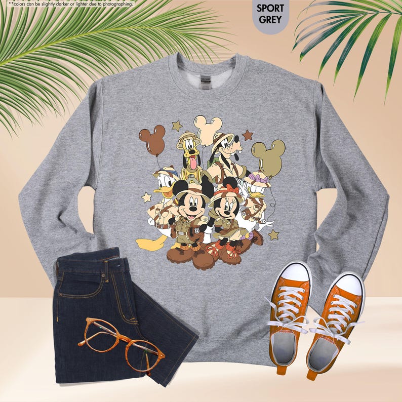 May include: A gray sweatshirt featuring a cartoon graphic of Mickey Mouse, Minnie Mouse, Donald Duck, Goofy, and Pluto in safari outfits. The sweatshirt is paired with dark blue jeans, orange-framed glasses, and orange sneakers.