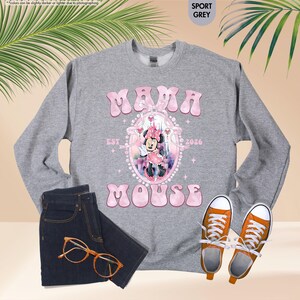 May include: A gray sweatshirt with the text "MAMA MOUSE EST 2026" in pink, featuring a Minnie Mouse graphic. The image includes dark blue jeans, orange-framed glasses, and orange sneakers with white laces.