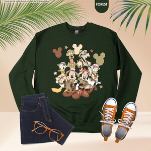 May include: Forest green sweatshirt featuring a graphic of Mickey Mouse, Minnie Mouse, Donald Duck, Goofy, and Pluto in safari outfits. The image also includes a pair of blue jeans, orange sneakers, and orange eyeglasses.