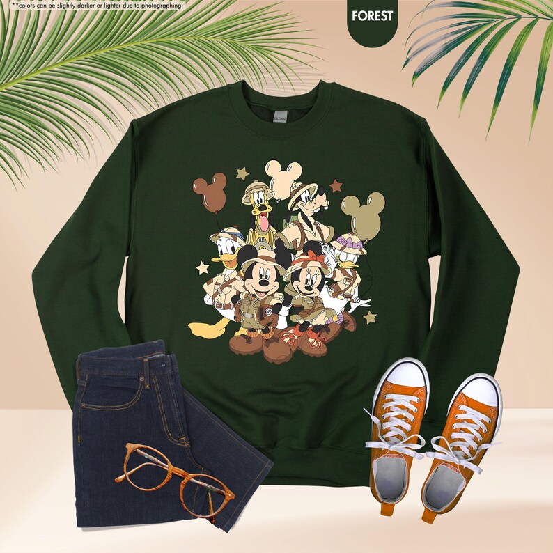 May include: Forest green sweatshirt featuring a graphic of Mickey Mouse, Minnie Mouse, Donald Duck, Goofy, and Pluto in safari outfits. The image also includes a pair of blue jeans, orange sneakers, and orange eyeglasses.