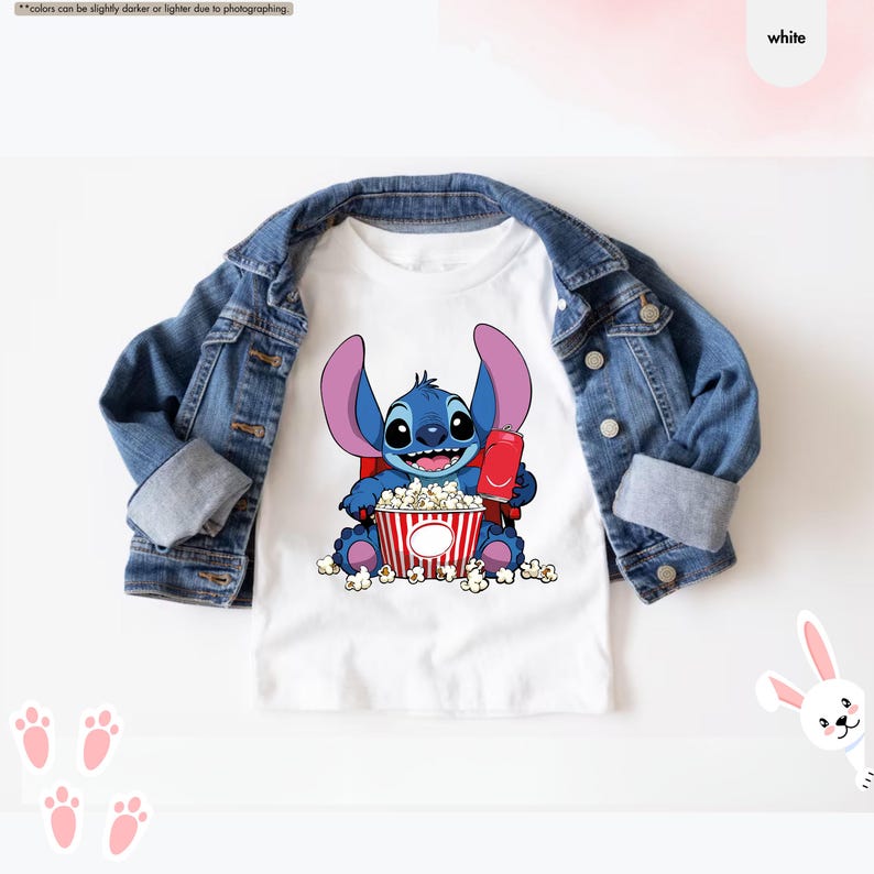 May include: White t-shirt featuring a blue cartoon character holding a popcorn bucket and a red drink. The shirt is paired with a denim jacket. The character has large ears and is surrounded by popcorn.