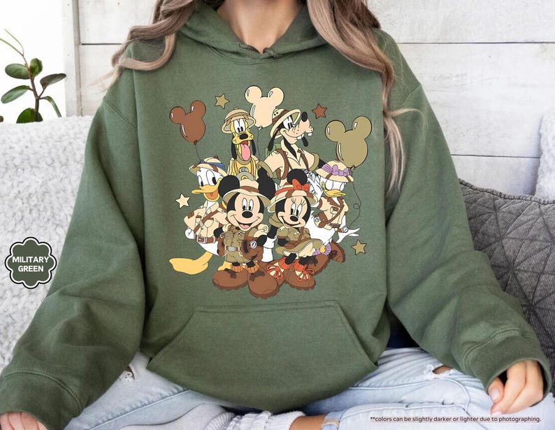 May include: Military green hoodie featuring a graphic of Mickey Mouse, Minnie Mouse, Donald Duck, Goofy, and Pluto in safari outfits. The design includes brown Mickey Mouse-shaped balloons and stars. The hoodie has a kangaroo pocket and a drawstring hood.