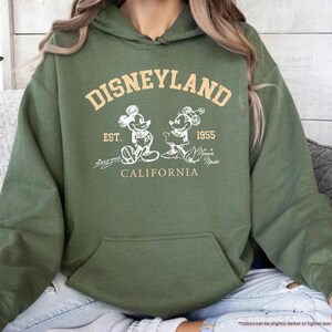 May include: Military green hoodie with a front pocket. The design features the text "DISNEYLAND" above an image of Mickey and Minnie Mouse, with "EST. 1955 CALIFORNIA" below. The hoodie is a casual style.