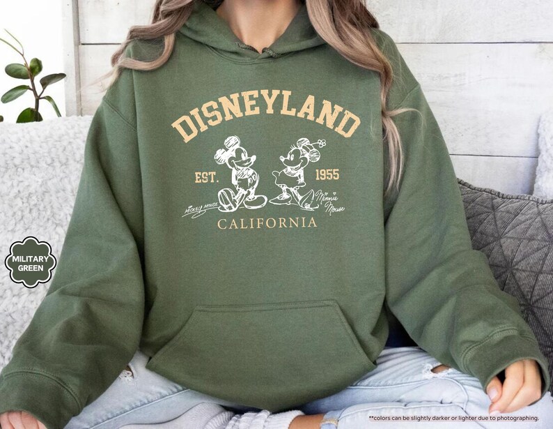 May include: Military green hoodie with a front pocket. The design features the text "DISNEYLAND" above an image of Mickey and Minnie Mouse, with "EST. 1955 CALIFORNIA" below. The hoodie is a casual style.