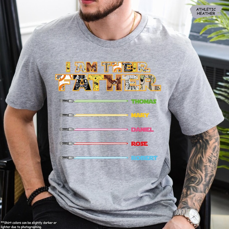 Puede incluir: Camiseta gris atl&eacute;tica jaspeada con el texto "I AM THEIR FATHER" en un dise&ntilde;o colorido. Debajo, ilustraciones de sables de luz con los nombres: Thomas, Mary, Daniel, Rose y Robert.