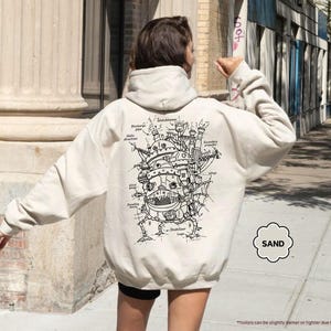 May include: A cream-colored hoodie with a black and white illustration of a flying machine on the back. The illustration includes text labels for different parts of the machine. The text "SAND" is in a circle on the bottom right corner of the hoodie.
