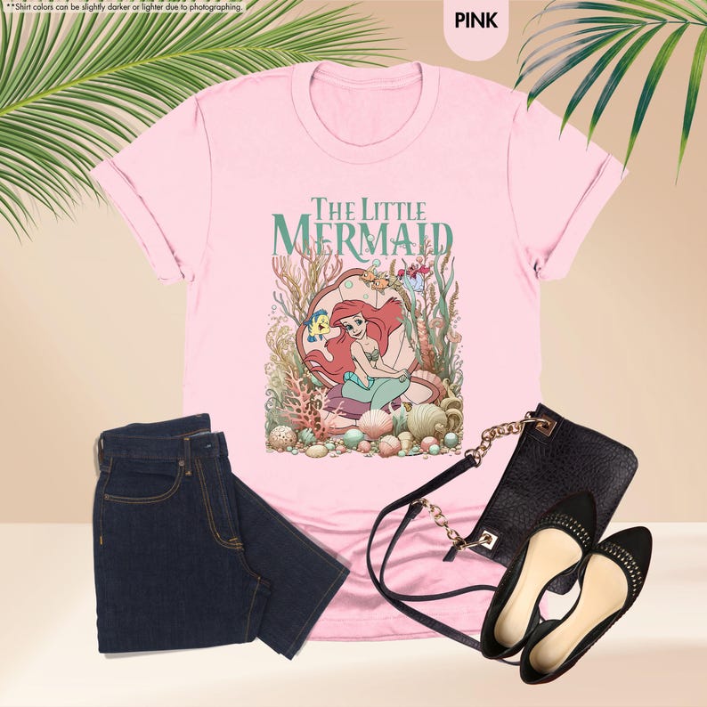 May include: Pink t-shirt featuring "The Little Mermaid" graphic, paired with dark blue jeans, a black purse, and black flats. The shirt has rolled-up sleeves and the graphic shows Ariel in a seashell.