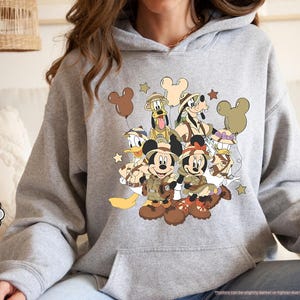 May include: Sport grey hoodie featuring a graphic of Mickey Mouse, Minnie Mouse, Donald Duck, Goofy, and Pluto in safari outfits. The design includes brown Mickey Mouse-shaped balloons and stars. The hoodie has a kangaroo pocket and a drawstring hood.