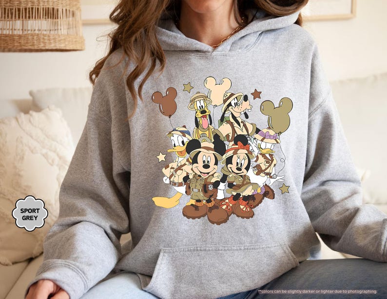 May include: Sport grey hoodie featuring a graphic of Mickey Mouse, Minnie Mouse, Donald Duck, Goofy, and Pluto in safari outfits. The design includes brown Mickey Mouse-shaped balloons and stars. The hoodie has a kangaroo pocket and a drawstring hood.