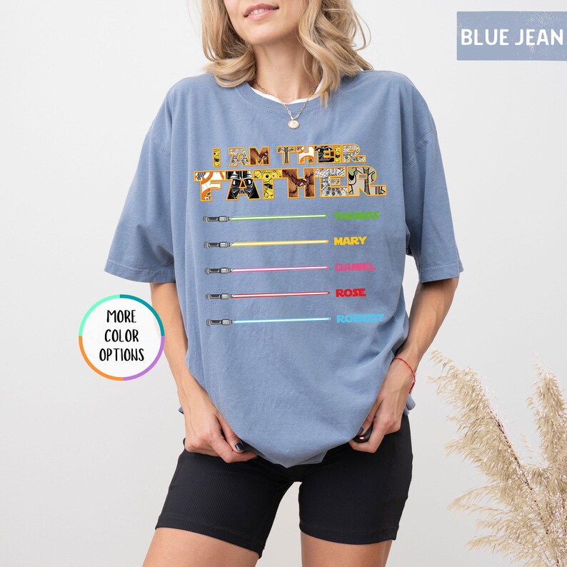 Puede incluir: Camiseta azul vaquero con el texto "I AM THE FATHER" en un dise&ntilde;o estampado. Debajo, gr&aacute;ficos de sables de luz con los nombres: Mary, Daniel, Rose y Robert. Un bot&oacute;n dice "MORE COLOR OPTIONS".
