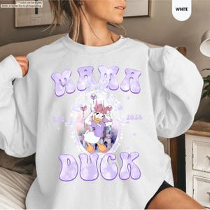 May include: White sweatshirt with the text "MAMA DUCK" in purple bubble letters. The design includes a cartoon image of Daisy Duck, a castle, and the year 2026. The sweatshirt is a casual, comfortable style.